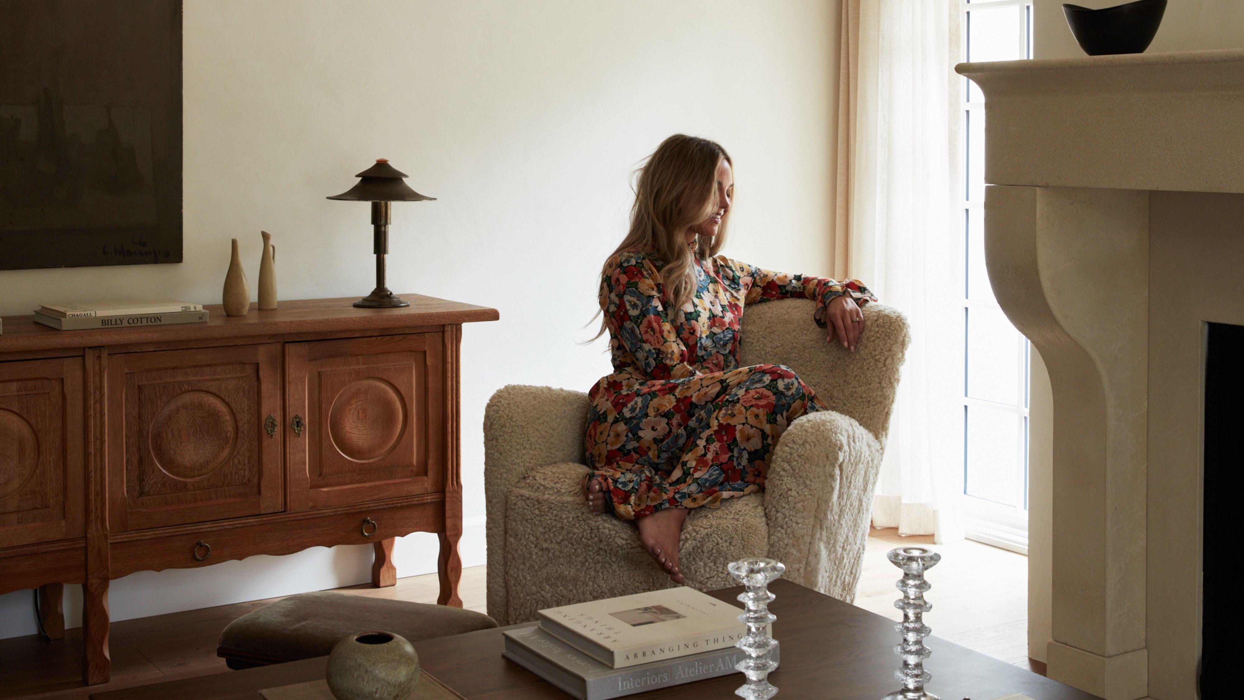 Interior designer Molly Kidd poses atop of a shearling accent chair by Maiden Home inside of her warm minimalist Oregon living room, which features collected wooden, brass, and ceramic objects.
