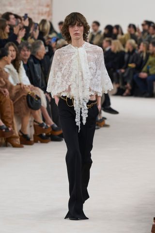 A model at the F/W 24 Chloé show wearing a white lace cape top with black trousers and a gold Chloé belt.