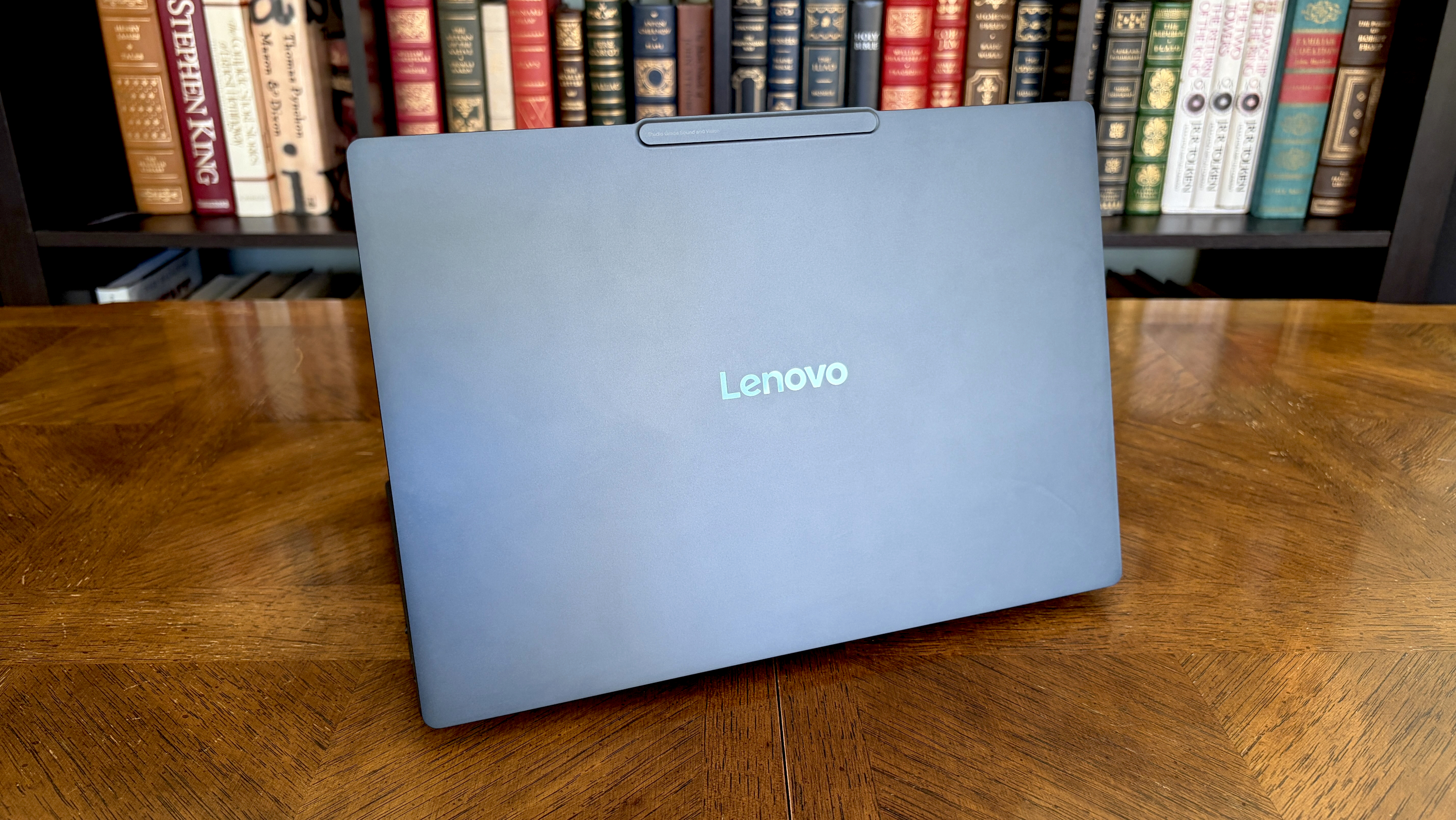 A closed Lenovo laptop with a dark, brushed-metal finish sits on a wooden table. Behind it, a bookshelf filled with colorful hardcover books.