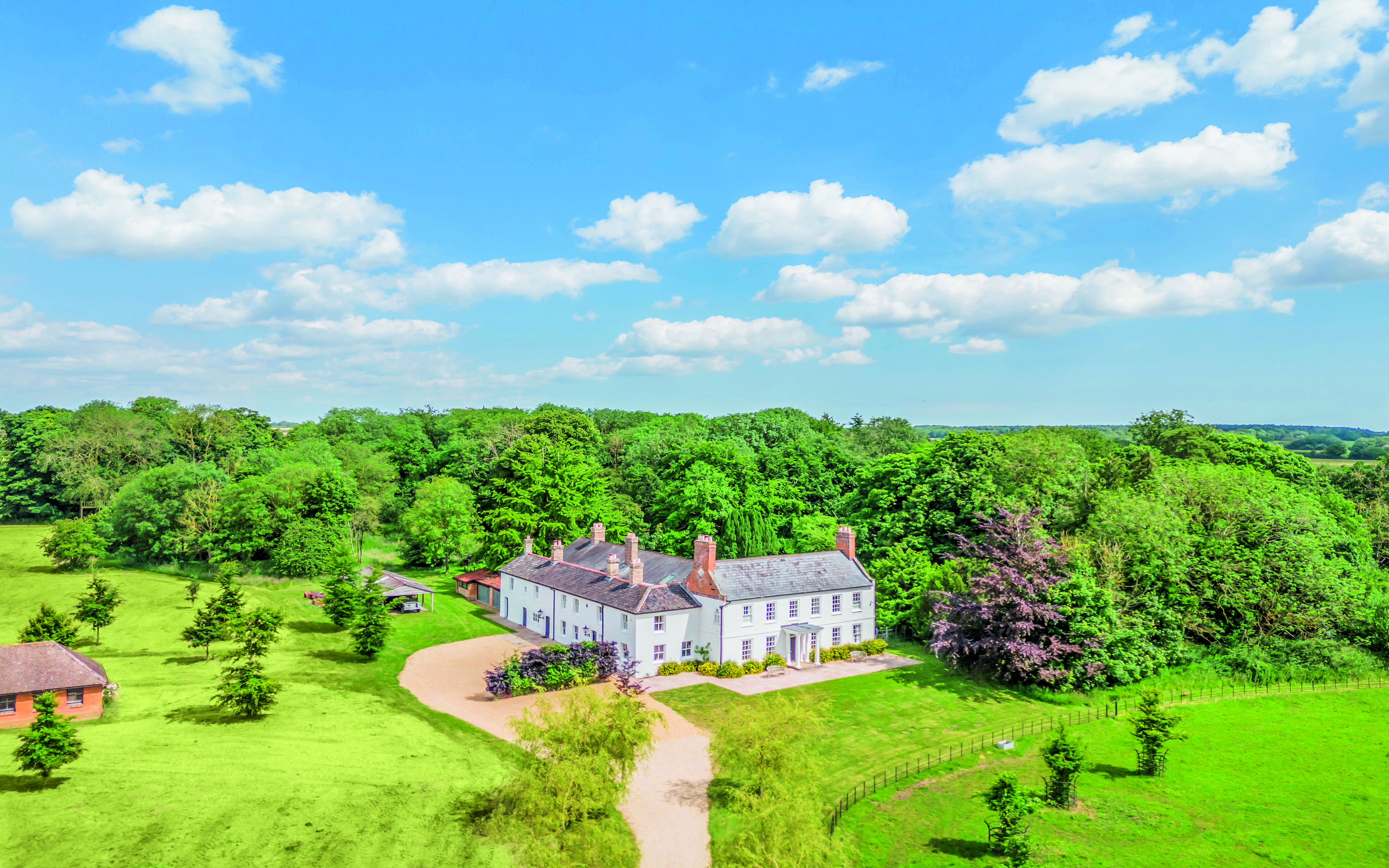 Exterior view of Saham Lodge, showing its two-storey elevation with a pitched roof, as well as its white exterior. It's surrounded by lush green countryside 