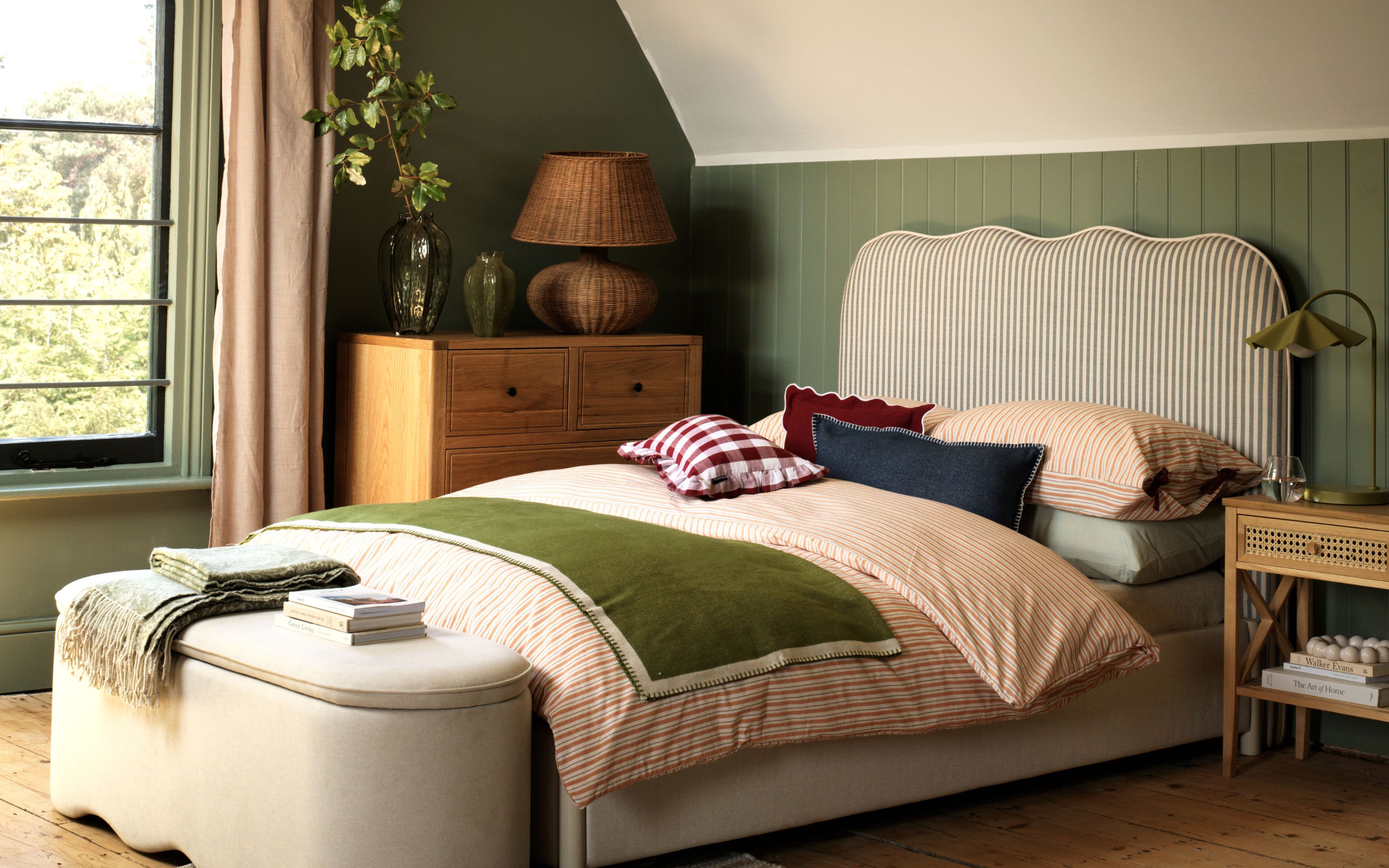 Green bedroom with wooden chest, rattan large lamp and bedside table. Striped headboard with red patterned bedding and green blanket. Natural ottoman at bottom of bed