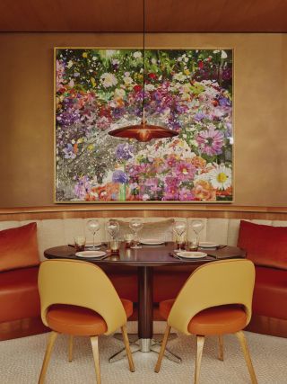 A 1970s-inspired dining room features leather and wood furniture, brass lighting, and a large floral painting on the walls.