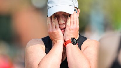 Top-half of a female runner shown with her hands on her face against a blurred background. She looks completely exhausted. She is wearing a black vest, red watch and white baseball cap, as well as blue nail varnish.