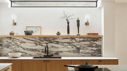A kitchen with a marble backsplash. There is a black sink with a black colored tap. Above the sink is open shelving with decor and wall lights. In the frame is also a stovetop and pan.