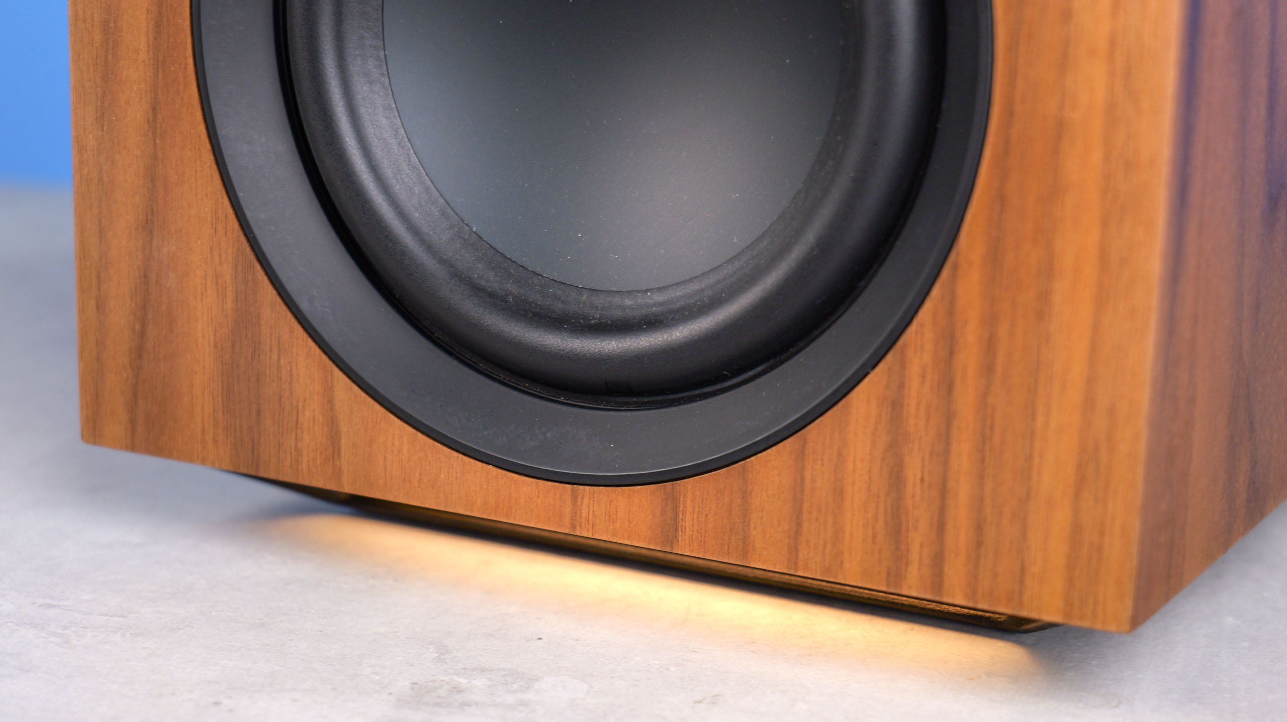 The Cambridge Audio L/R X speakers underlight, in walnut color, positioned on a stone surface with a blue background