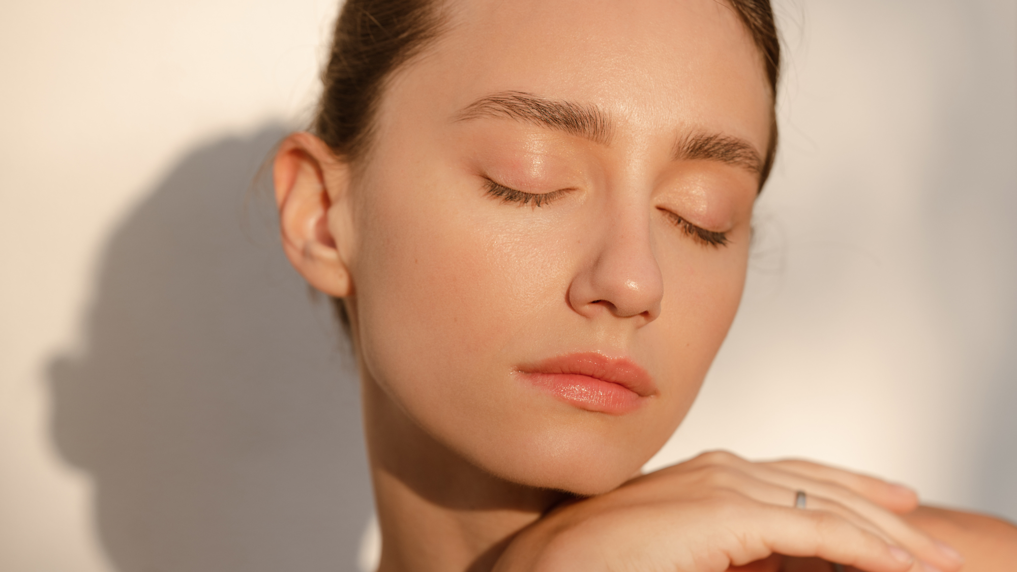Woman standing against a white wall closing her eyes with the sun beaming down on her