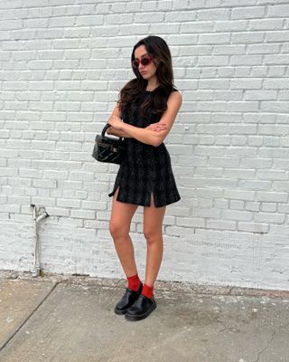 Woman wearing black dress, shoe clog trend, and red socks leaning against wall.