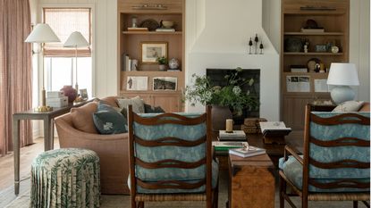 farmhouse meets grandma chic aesthetic white living room with tongue and groove walls, light oak built in shelving, fireplace, soft pink couch, skirted floral ottoman and two dark wood armchairs with blue cushions