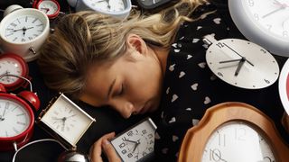 A woman with blonde hair sleeps surrounded by alarm clocks of different colours
