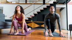 A woman and man dressed in leggings and workout tops on exercise mats next to each other facing the camera and performing a deep lunge looking upward. there's a modern living room background with stairs and mezzanine floor. 