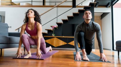 A woman and man dressed in leggings and workout tops on exercise mats next to each other facing the camera and performing a deep lunge looking upward. there's a modern living room background with stairs and mezzanine floor.