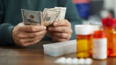 Close-up of a person's hands counting dollars near prescription pill bottles and a pill organizer.