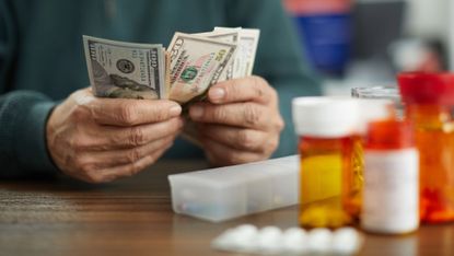 Close-up of a person's hands counting dollars near prescription pill bottles and a pill organizer.