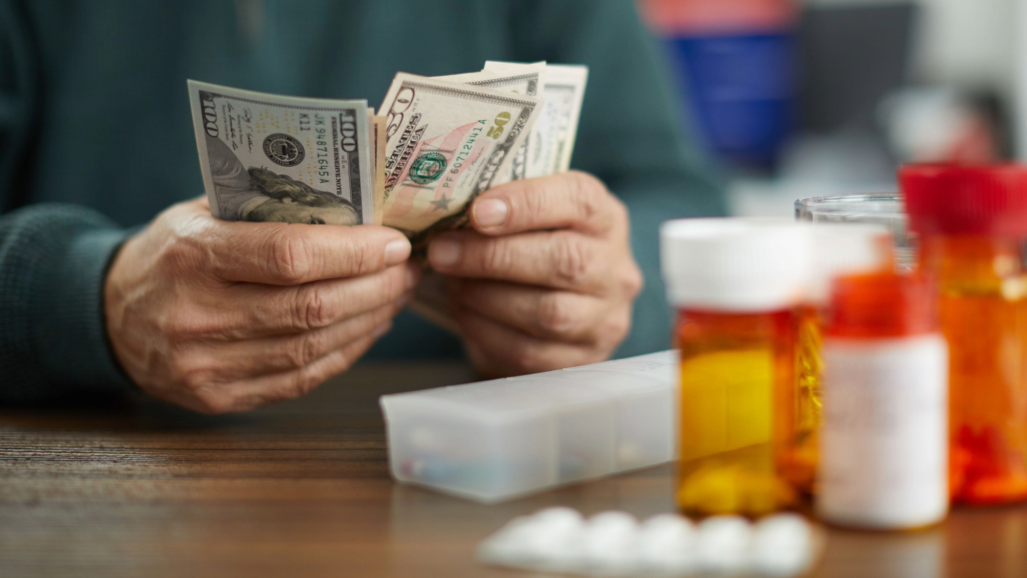 Close-up of a person's hands counting dollars near prescription pill bottles and a pill organizer.