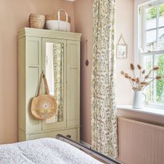 pink bedroom with a cream wardrobe in the corner, and a wicker bag hanging from the handle