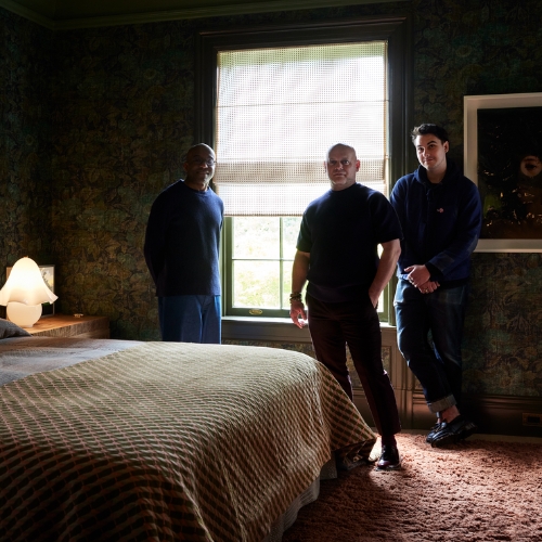 Image of three men standing by a window in a moody bedroom.