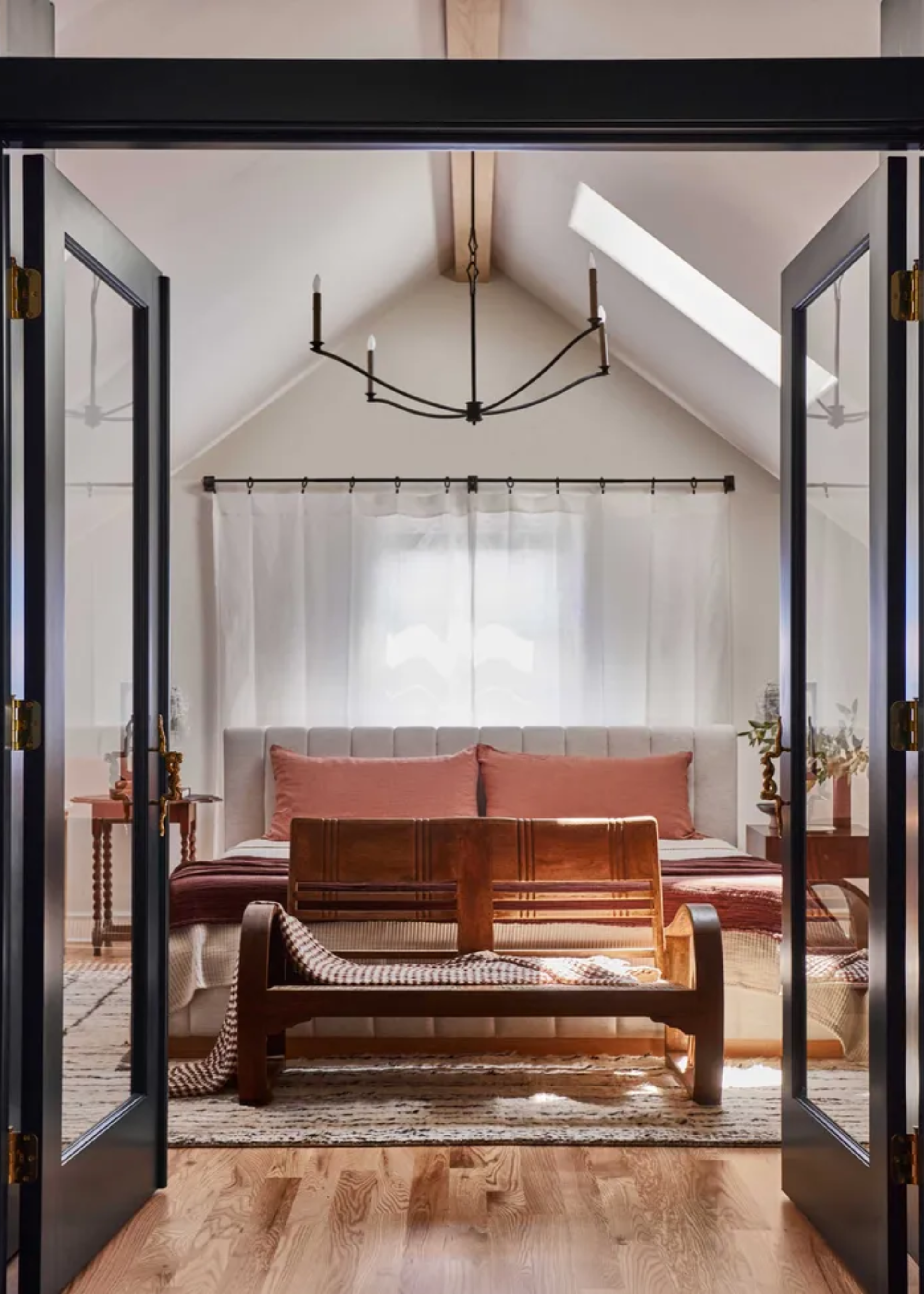 A bedroom with a high ceiling, a hanging iron candelabra chandelier, a bed with pink pillows, bobbin-ed side tables, a bench, and a textured area rug