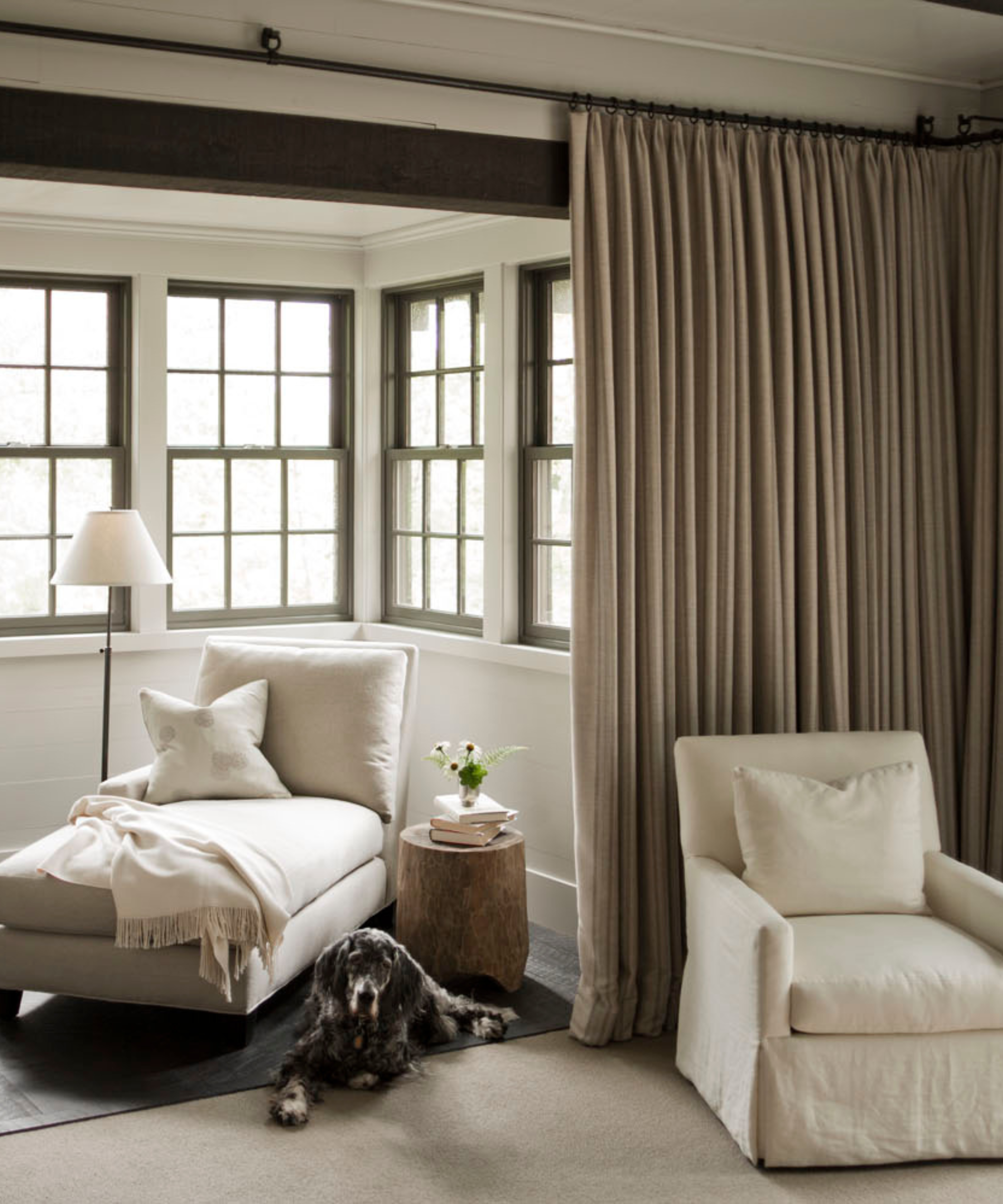 a sitting area in a bedroom with long taupe drapery, a white chaise lounge, a white accent chair, and a dog sitting on the floor