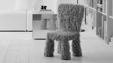 black and white image of furry chair in living room in front of mirrored side table, white sofa, and bookcase with white floorboards