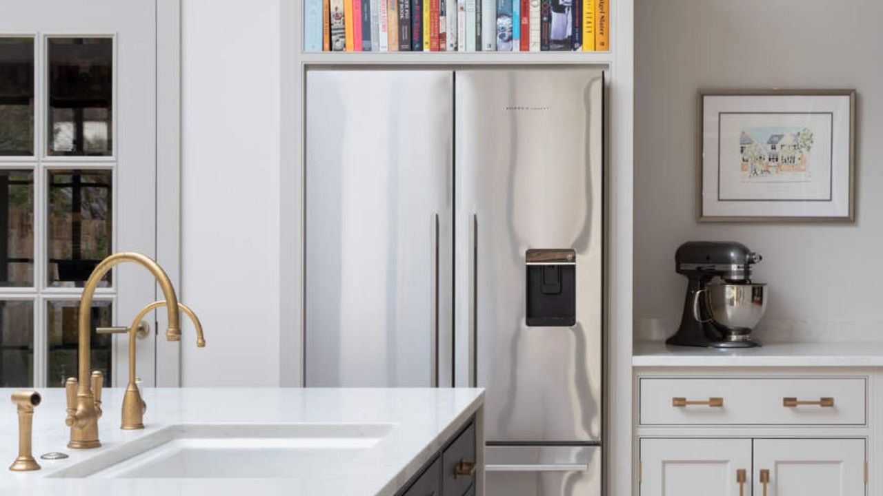 A silver double door fridge in a white kitchen. Nestled in built in cabinets, cookbook storage on top , a gold tap sink in the foreground