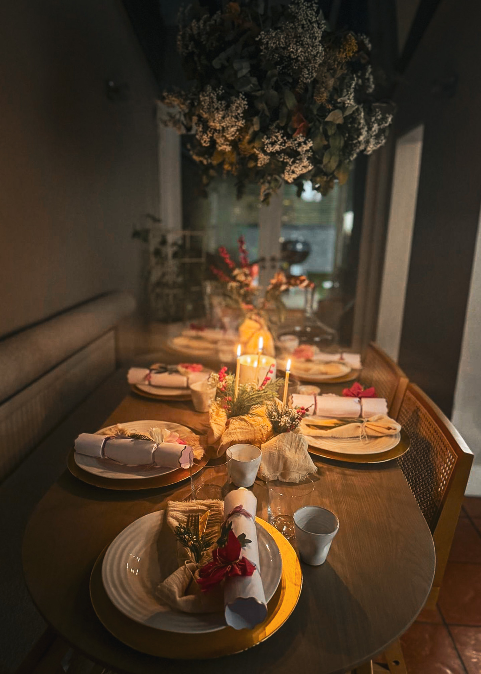 A dining table with banquette seating on one side and chairs on the other is decorated for christmas with lit candles, crackers on each place setting and a floral and foliage display hangs above the table