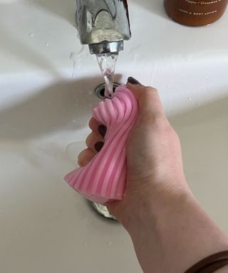 A woman's hand squeezing a pink damp duster under a running silver tap into a white bathroom sink