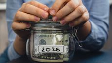 A pair of hands holding a glass jar full of American dollars that is labeled "2026"