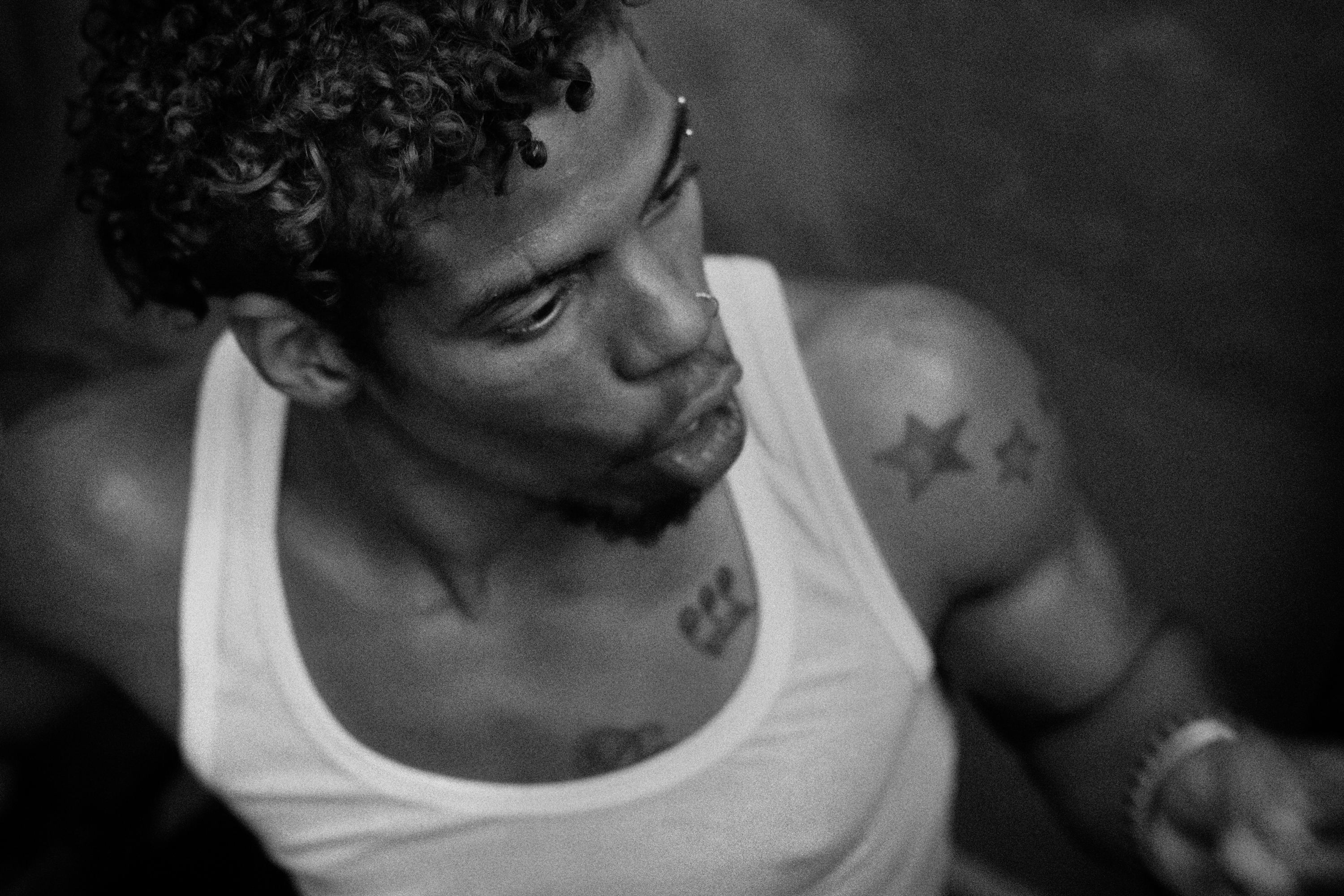 Black and white close-up of a young man in a white vest looking down, his neck and chest marked with star and crown tattoos and a small hoop earring visible.