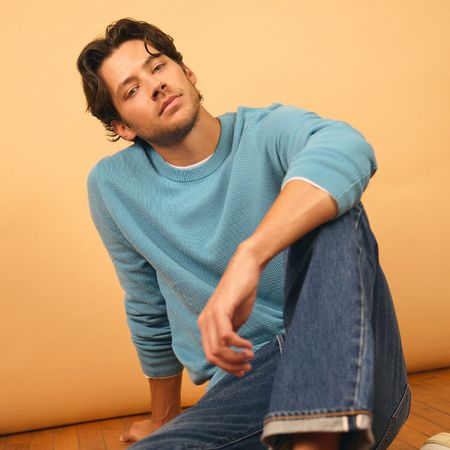 Matt Cornett photographed by Henry Wu and styled by Ashley Weston. He is sitting on a wood floor, his arm resting on his knee wearing a lightly blue crewneck sweater and blue jeans. 