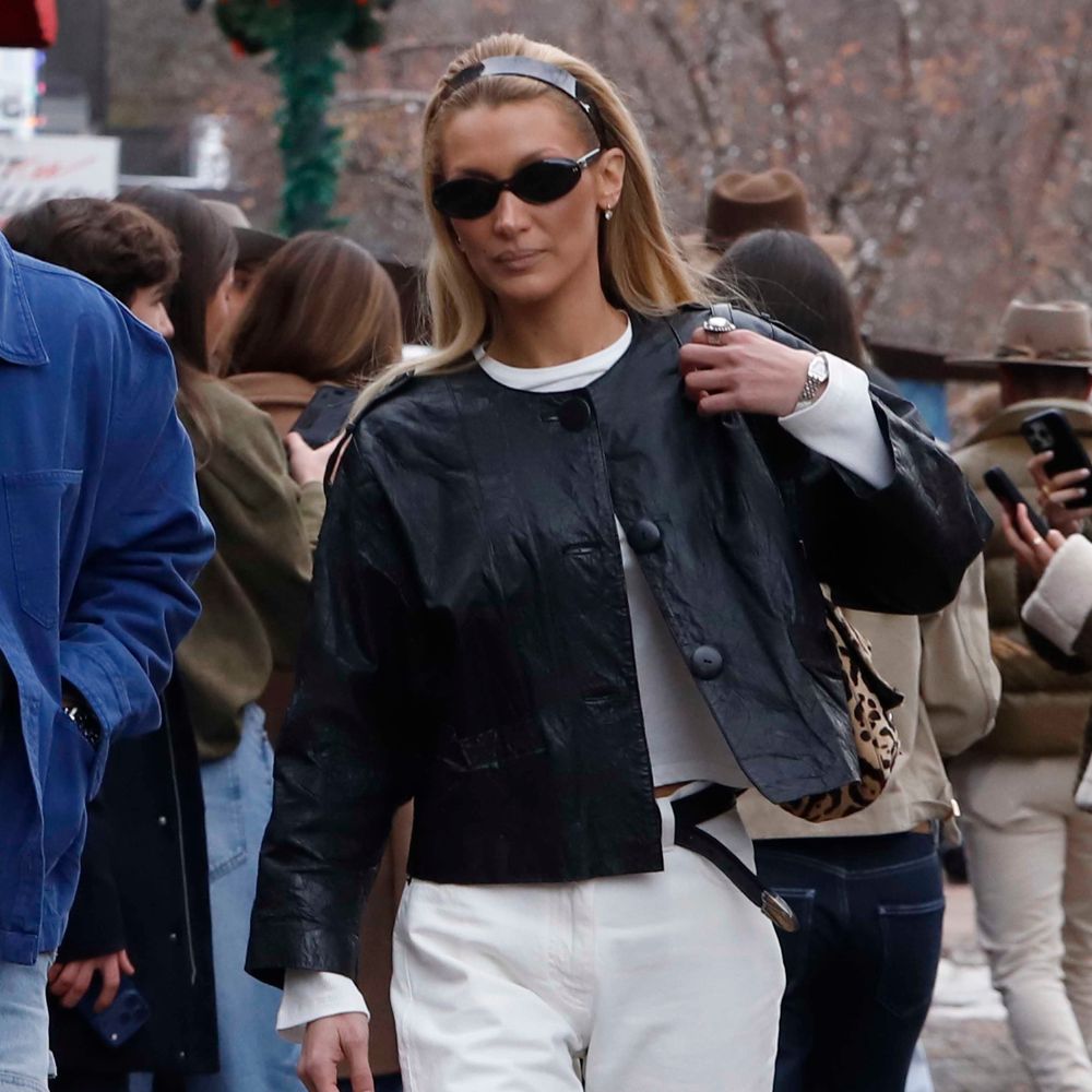 Bella Hadid walks along the pavement outside wearing white jeans with black boots and a black leather jacket.