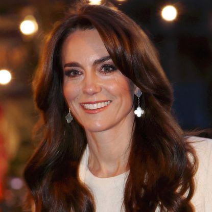 Kate Middleton wears silver earrings and a white sweater while wearing her long brown hair down and wavy