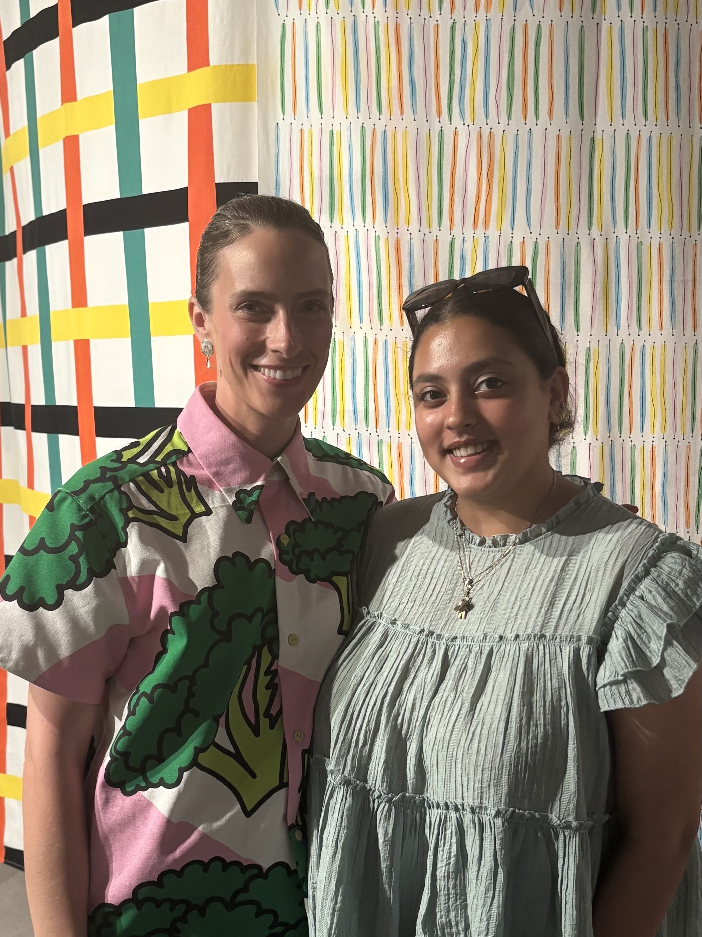 Two young women dressed in patterned shirts and denim jeans stand against a colorful textile installation made of stripy tents in green, orange, yellow, white, and black tones.