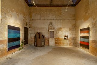 View of the 'Celtique' exhibition by Sean Scully at Château de Boisgeloup - multicoloured paintings by Scully in a space with distressed walls, a wooden door, a statue on a plinth against the wall and a wooden unit