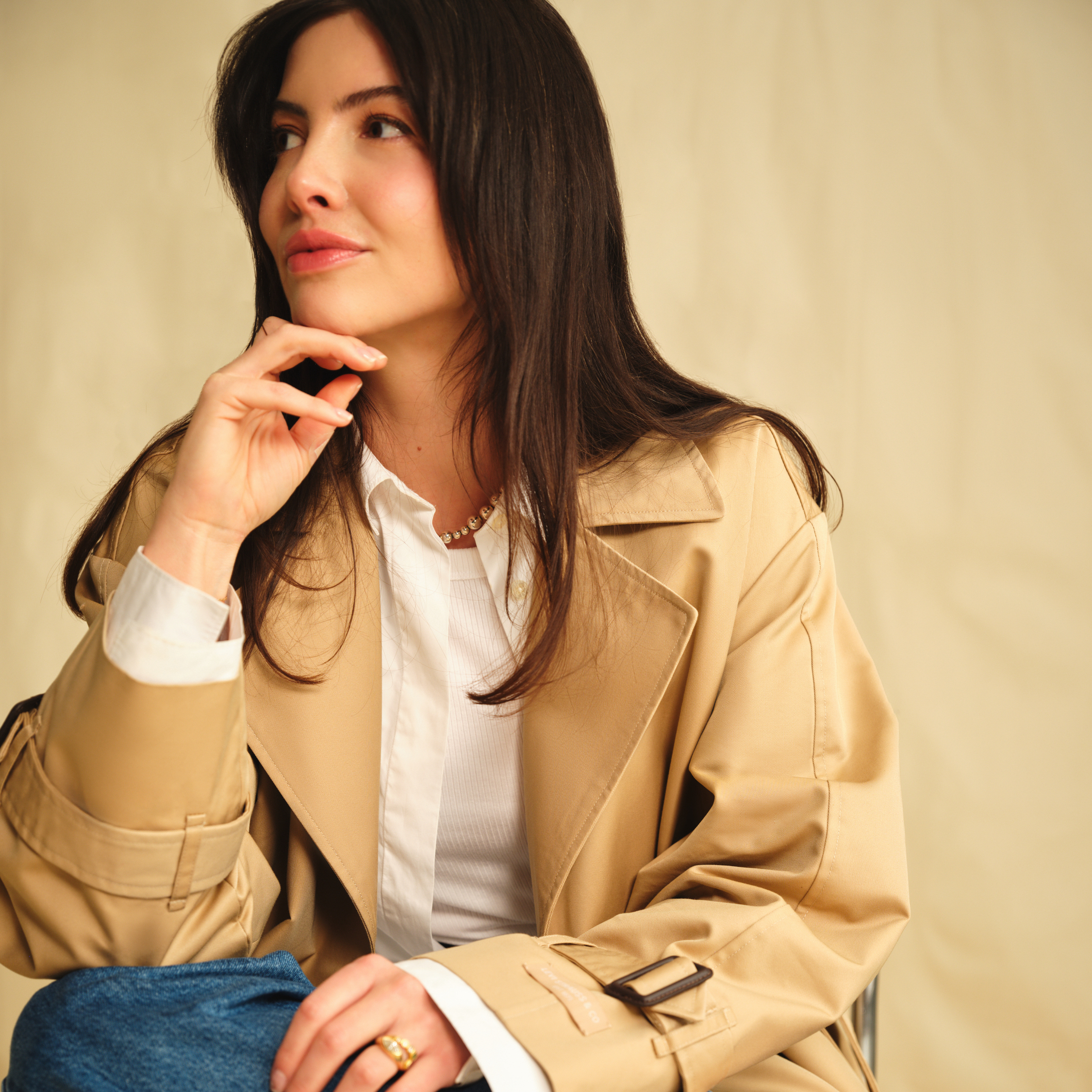 Kat Collings sitting with hand on chin wearing white collared shirt, tan trench coat, and jeans