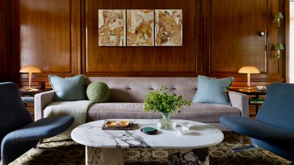 Living room with rich wood-panelled walls, lilac low-slung sofa and cool dark blue armchairs with marble coffee table