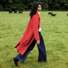 Model walking through a field with black sheep wearing wide leg dark denim trousers and long red coat for Nobody's Child discount codes