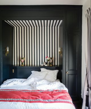 bedroom with black alcove with bed in the middle, striped black and white wallpapered accent wall, white bedding with pink throw