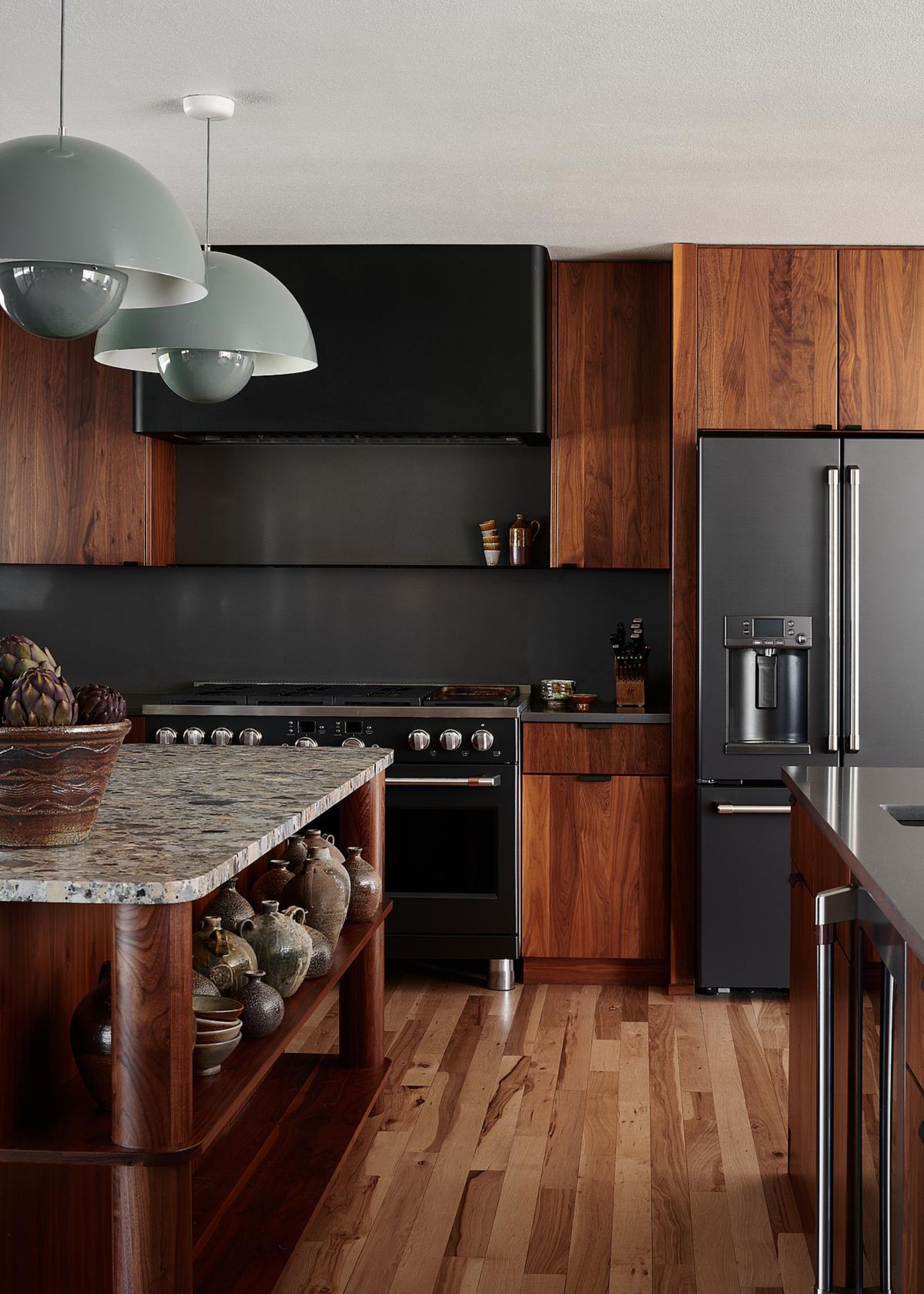 A dark wood kitchen with a large island in the middle and open shelving. There are also large pendant lights hanging above the kitchen island. You can also see a view of a large double door fridge as well as a stove.