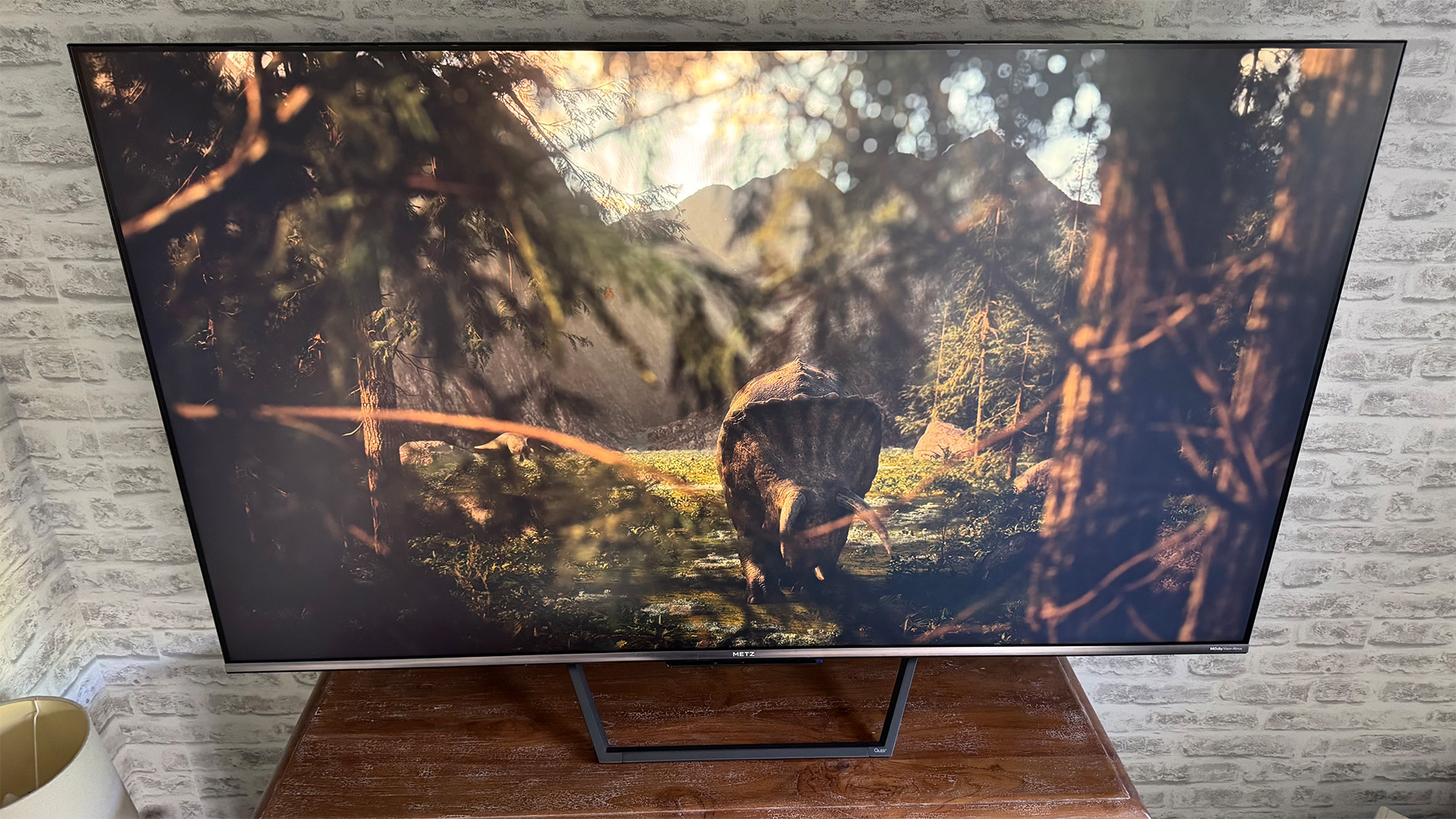 Metz MQE7600 4K TV on wooden surface in front of brick wall, on screen is triceratops