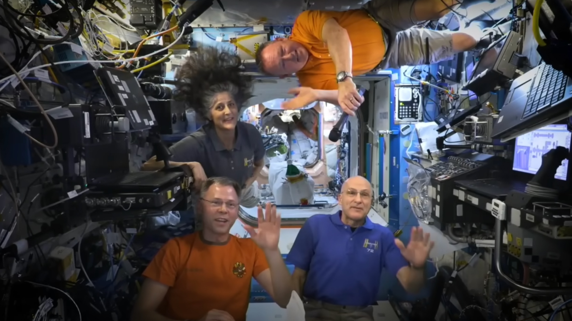 three men and one woman wave from inside the international space station