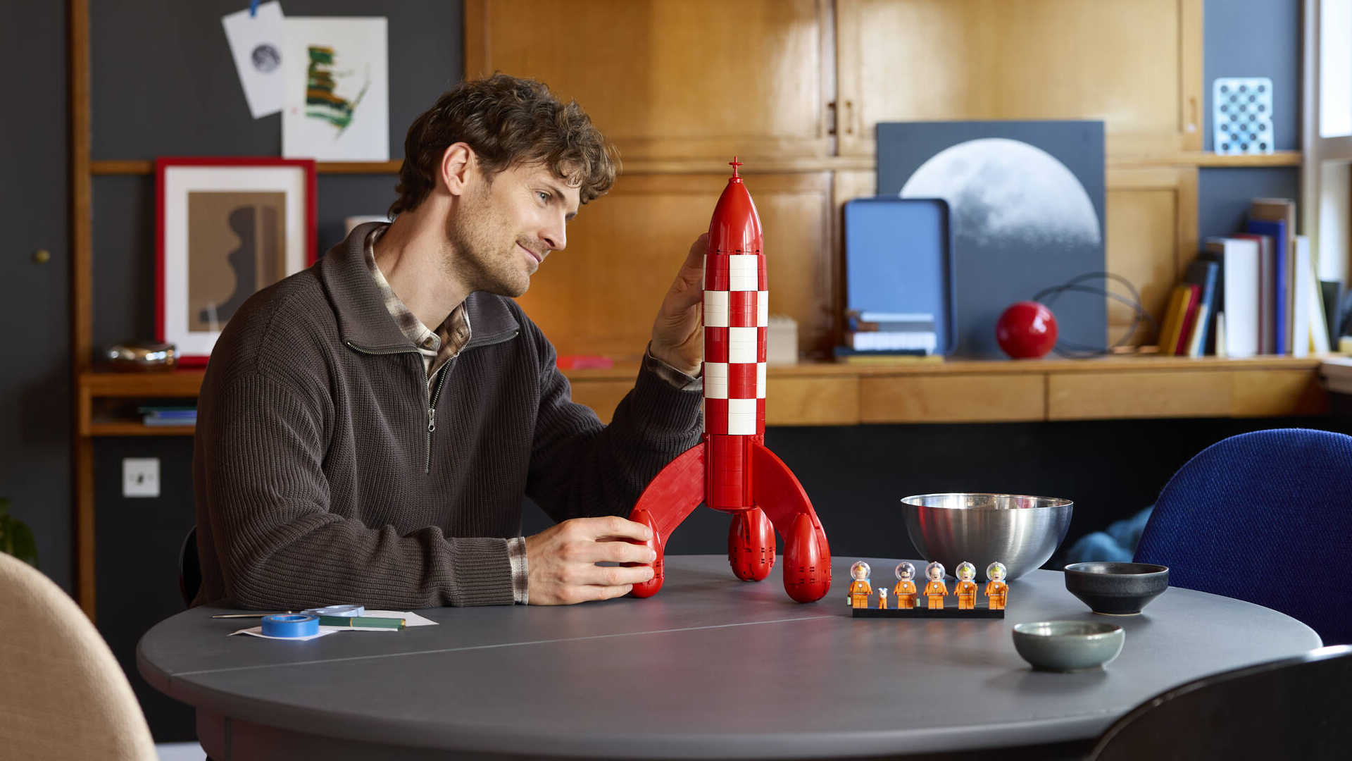A man places a piece on the Tintin Moon Rocket in a home environment, while smiling