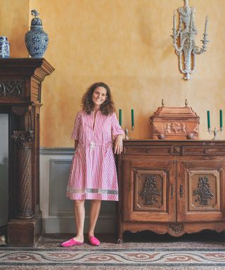 French house with authentic yellow walls, large antique sideboard and fire place, mosaic tiled floor, pottery, candlesticks, wall light, owner stood in pink stripe dress, pink shoes
