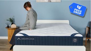 The DreamCloud Classic Hybrid Mattress on a bed frame in our sleep studio, a Tom's Guide Price Drop deals graphic in the bottom right foreground. A woman in a striped shirt sits on the bed's edge. 