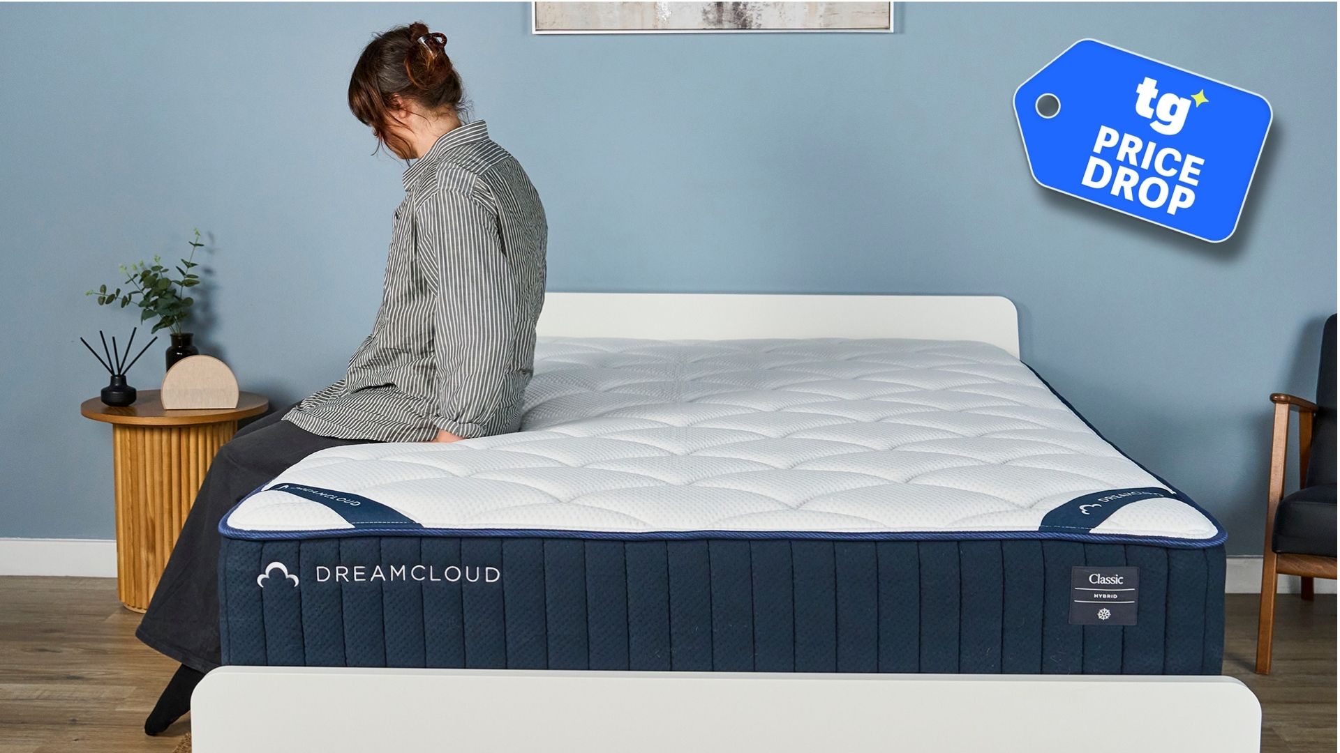 The DreamCloud Classic Hybrid Mattress on a bed frame in our sleep studio, a Tom's Guide Price Drop deals graphic in the bottom right foreground. A woman in a striped shirt sits on the bed's edge. 
