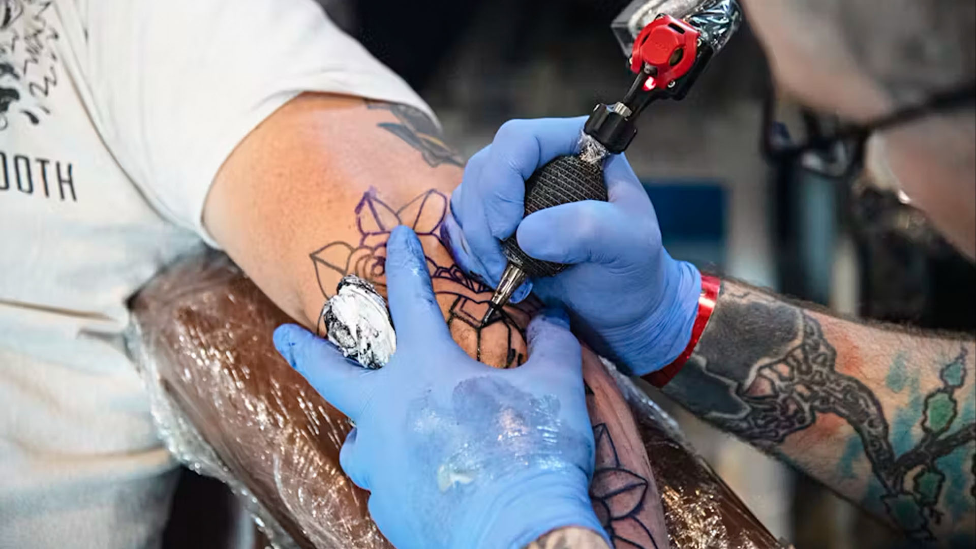 photo of a gloved tattoo artists using a tattooing gun to trace a design on a person's exposed arm