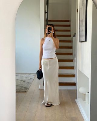 Influencer gaya Inggris Orlaith Melia berpose untuk selfie cermin di depan tangga mengenakan tank top putih berleher tinggi, rok slip berwarna gading, dan sandal flip-flop hitam.