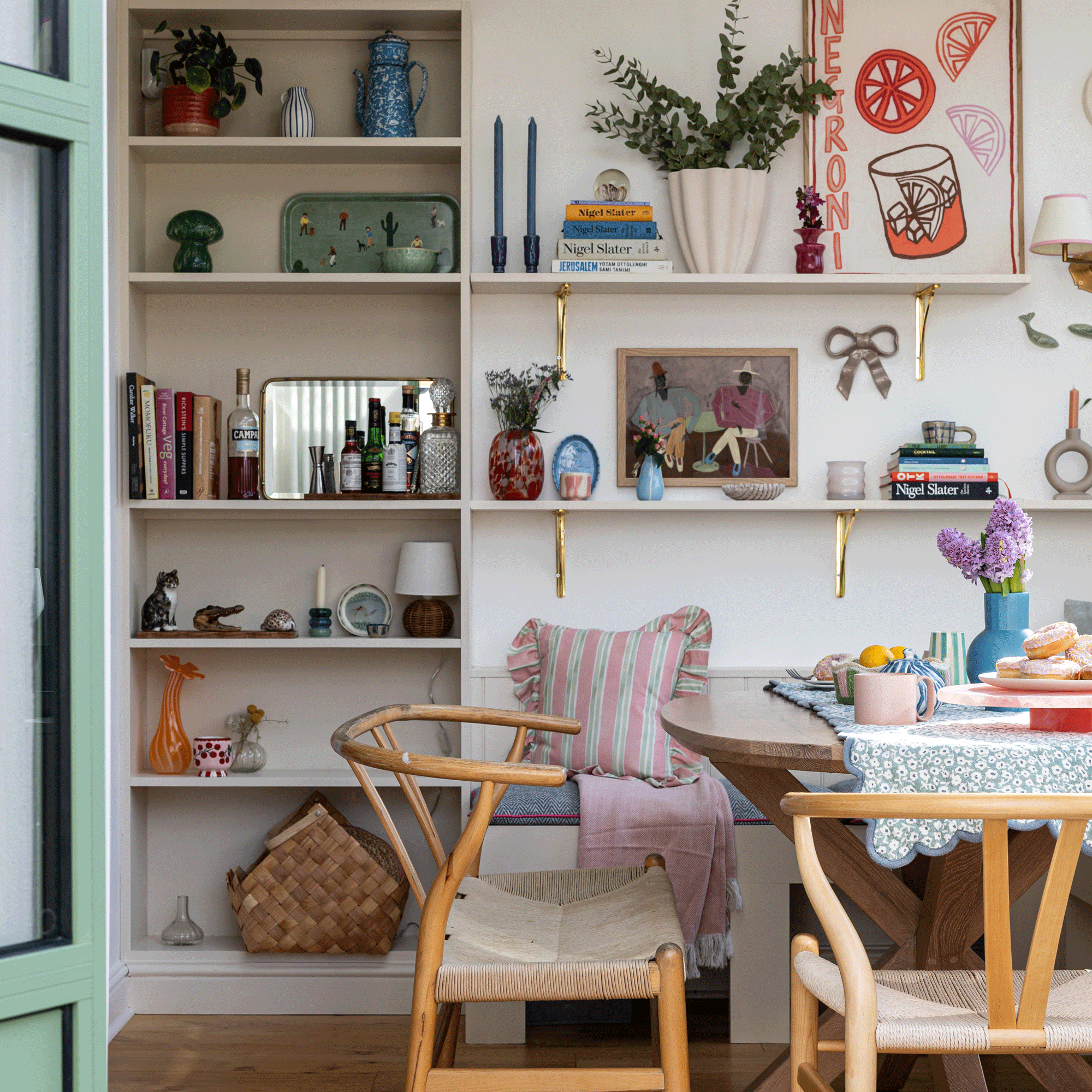 a dining room with a dining table and wishbone chairs, beside open shelving filled with books, art and accessories