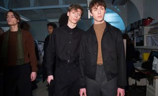 Four male models wearing looks from Margaret Howell's collection. One model is wearing a brown jumper, black trousers and green scarf. Another model is wearing a black shirt and trousers. Next to him is another model wearing a brown jumper, grey trousers and black jacket. And there is a fourth model in the background wearing a black coat