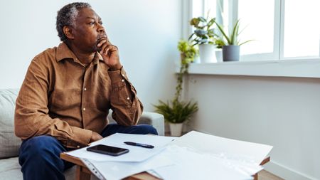 An older man sits in front of paperwork on his table and stares out the window, looking concerned.