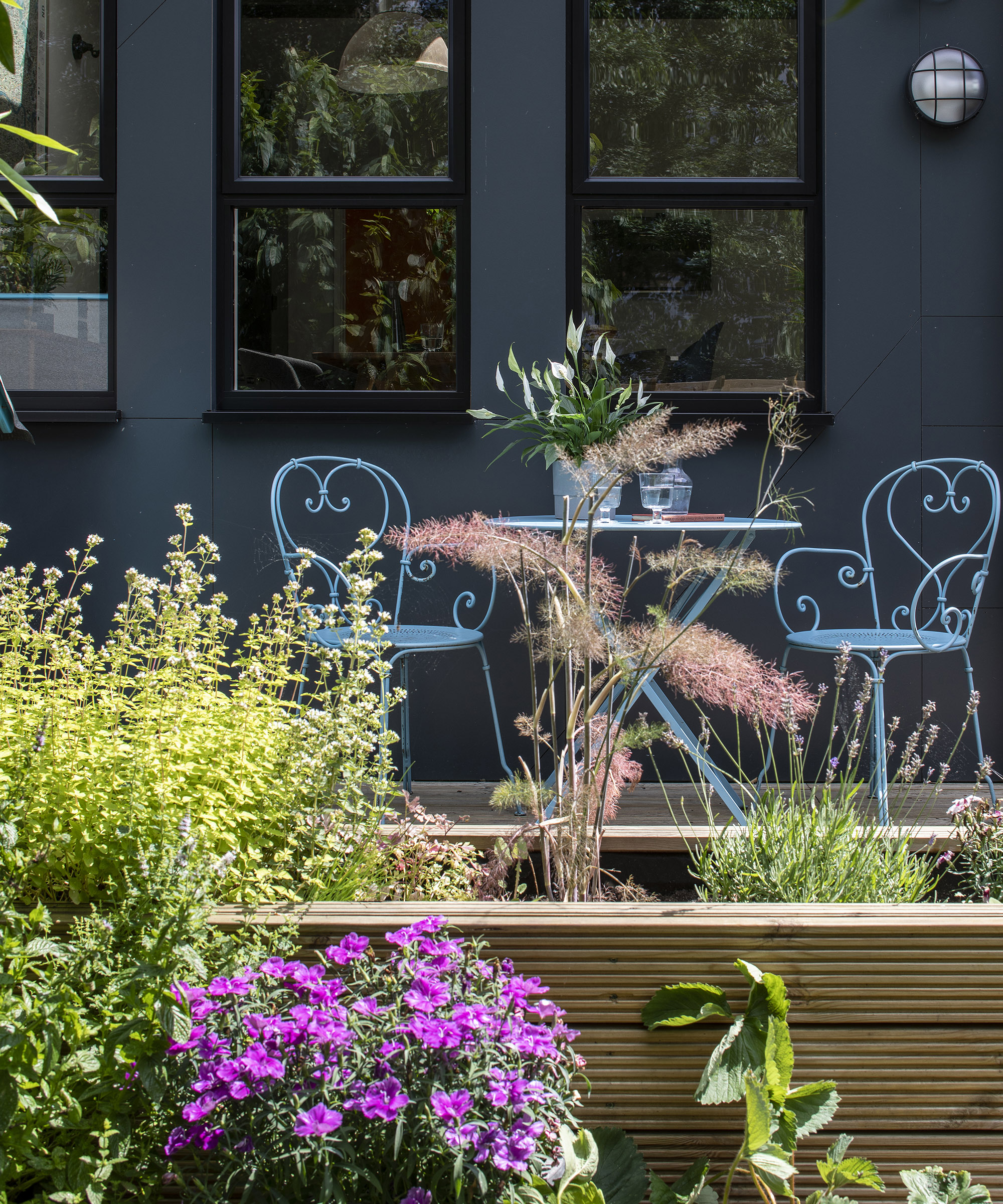 An outdoor bistro dining set on a patio outside a home extension with a timber raised bed in the foreground filled with planting